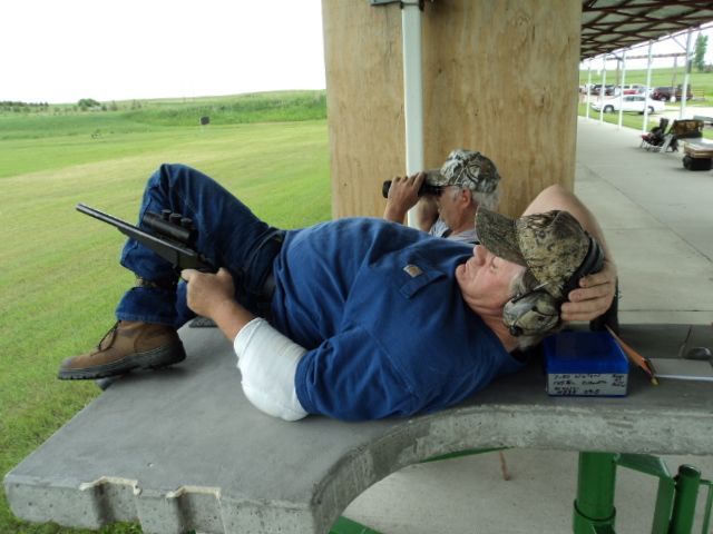 Man reclined on a shooting bench, holding a rifle; another man watches in the background at an outdoor range.