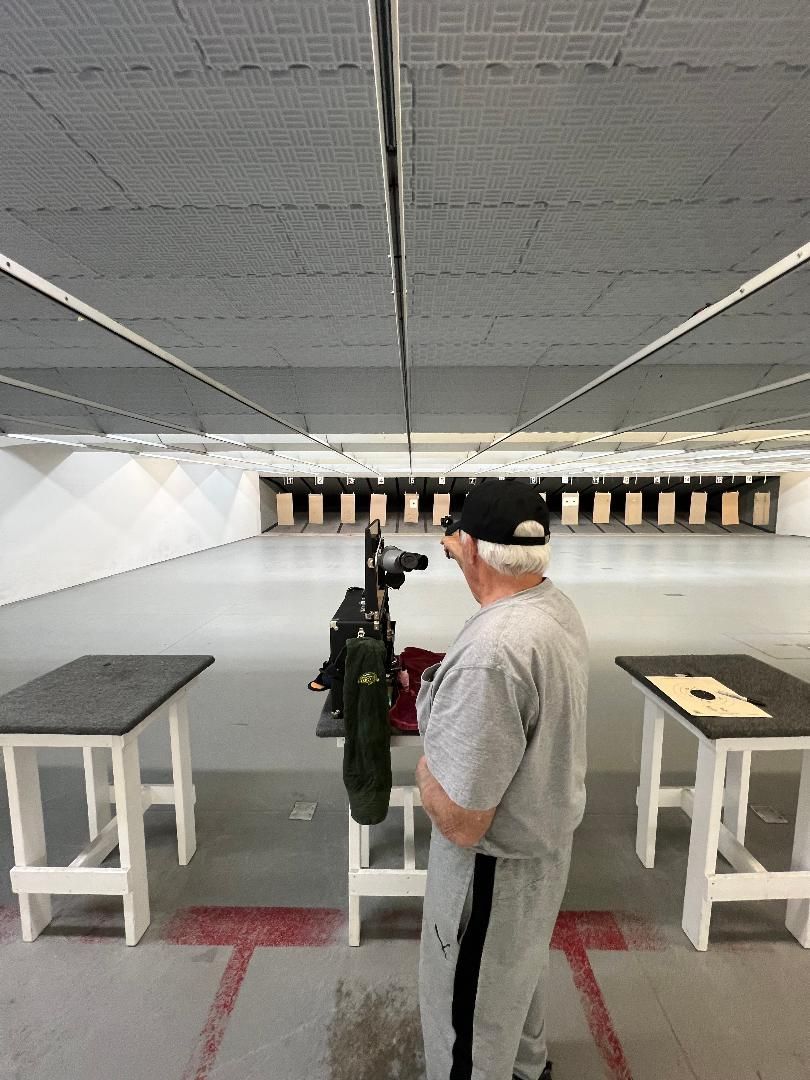 Man at a shooting range, aiming a rifle at targets. Wearing a cap and grey sweats.