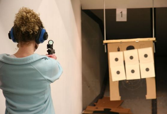 Woman at shooting range, aiming a handgun at target with multiple holes. She wears ear protection.