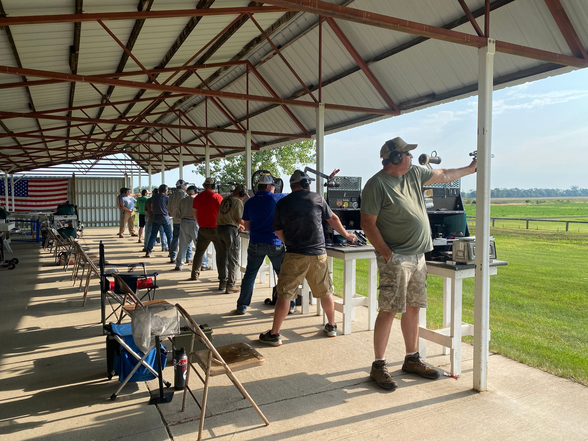 People shooting firearms at an outdoor range under a covered structure; American flag in background.