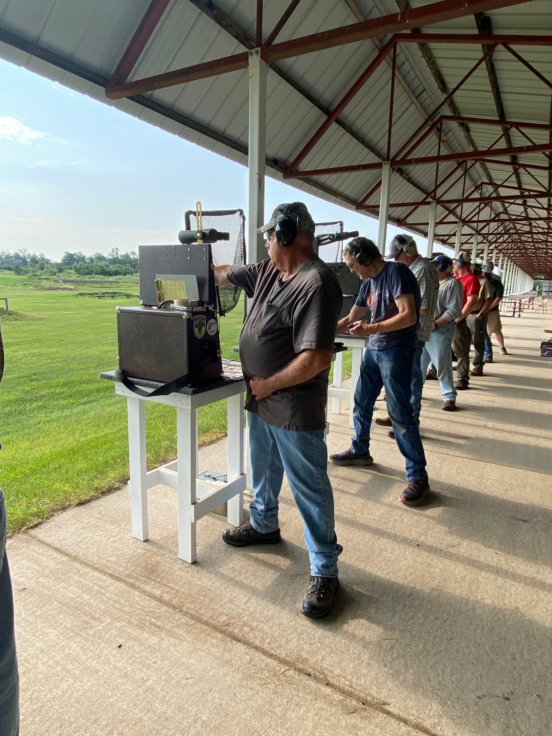 People shooting at a gun range. One man aims a gun at a target, others behind him. Outdoors, sunny day.
