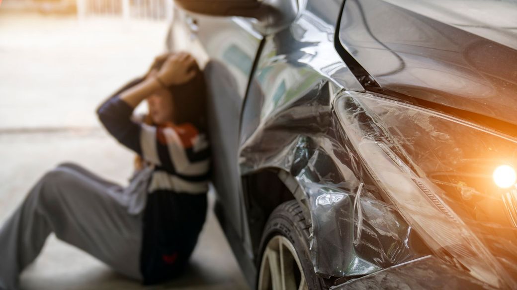 Person clutching head next to a damaged car, likely after an accident.