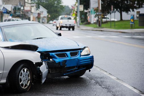 Two cars damaged in a collision on a wet city street; a blue car and a silver car.