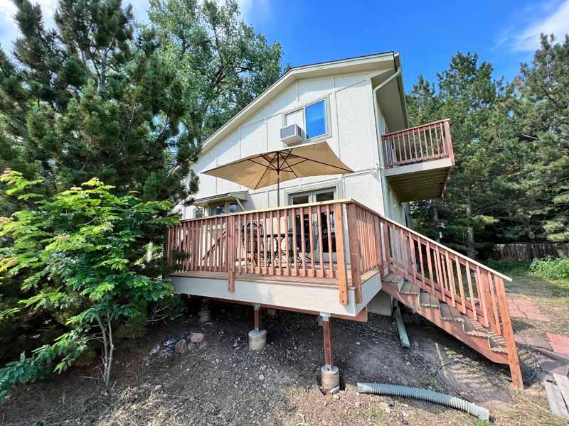 A two-story light-colored house featuring a wooden deck with stairs, a patio umbrella, and a small upper balcony.