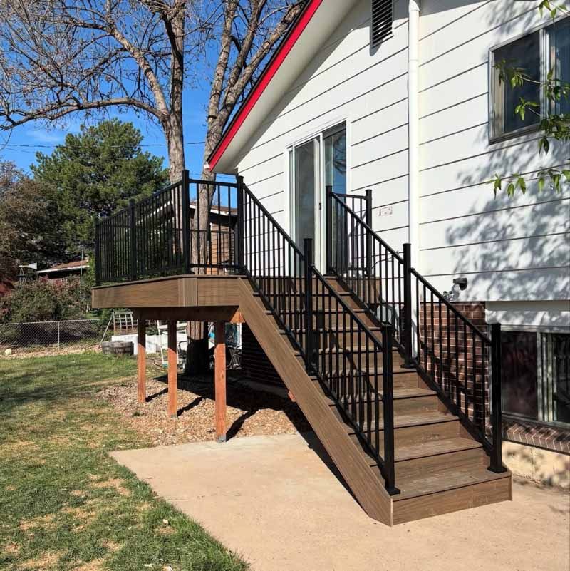 A wooden deck and staircase with black metal railings attached to the side of a white house leading to a backyard patio.