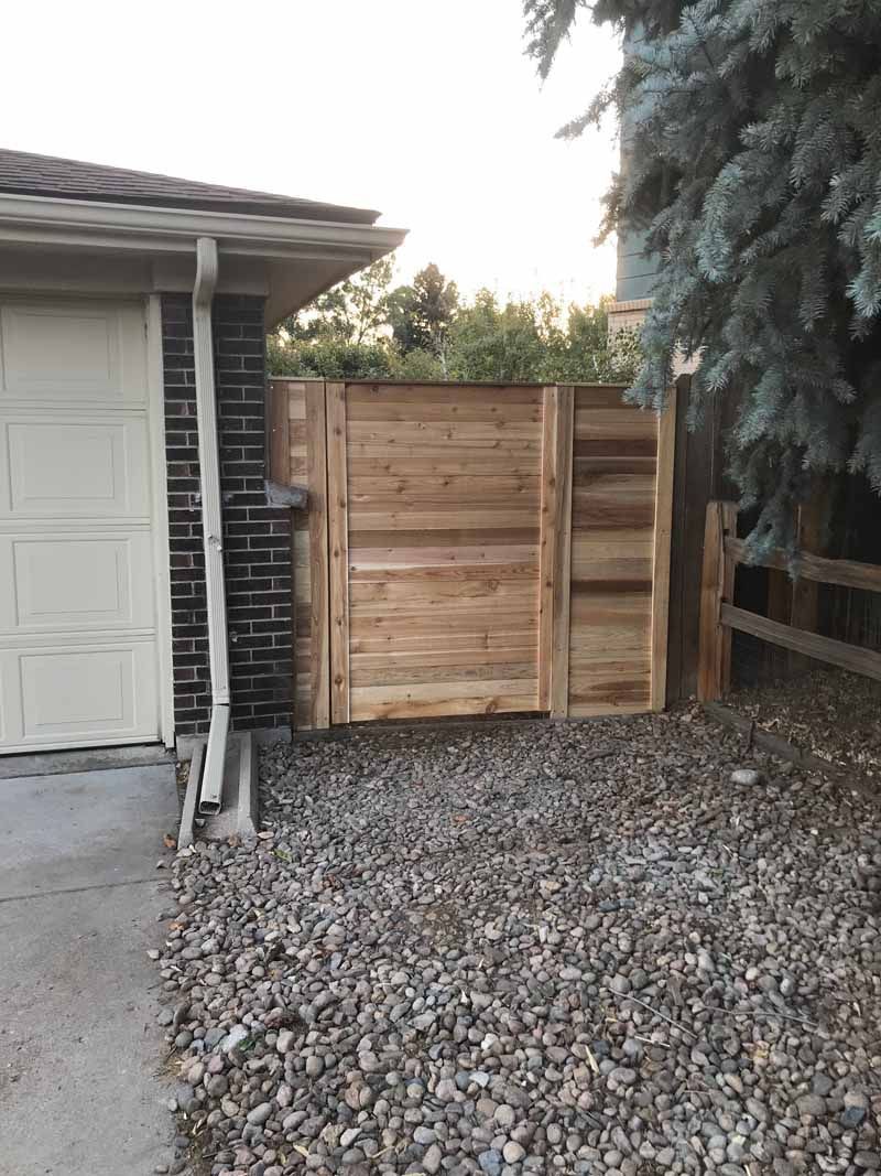 A wooden privacy fence section installed next to a garage wall, with a gravel yard in the foreground.