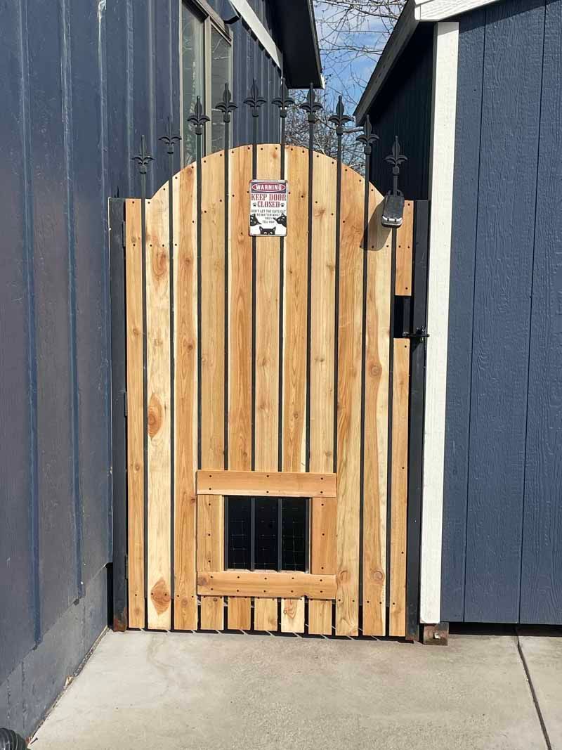 A wooden gate with vertical slats and black metal spikes at the top, featuring a small square viewing cutout with bars.