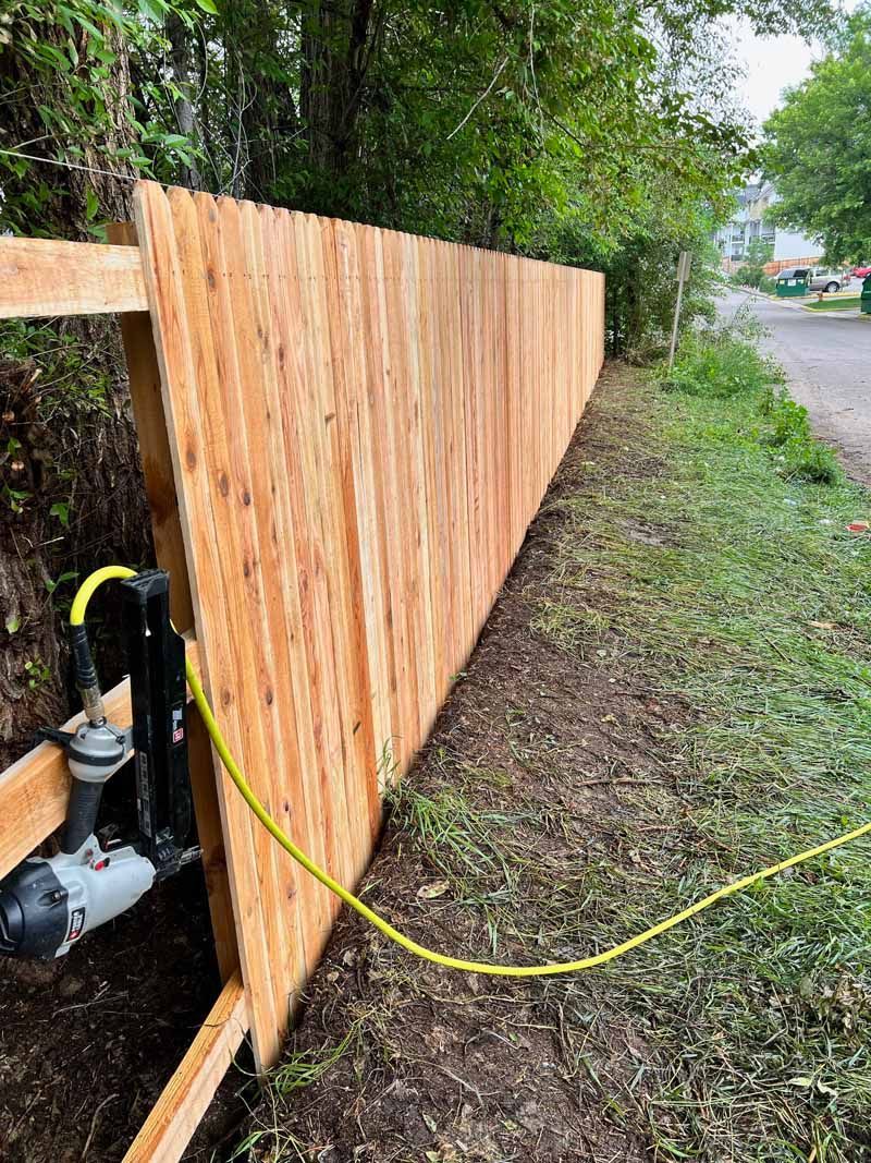 A pneumatic nail gun sits on a wooden frame against a newly constructed plywood fence outdoors.