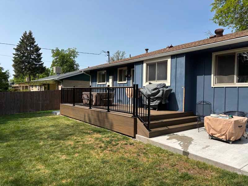 A deck with a black railing, wooden steps, and patio furniture attached to the rear of a blue house in a grassy yard.