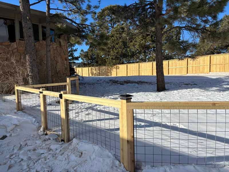 A wooden fence with wire mesh panels stands in a snowy yard, next to a stone-walled house and a tall evergreen tree.