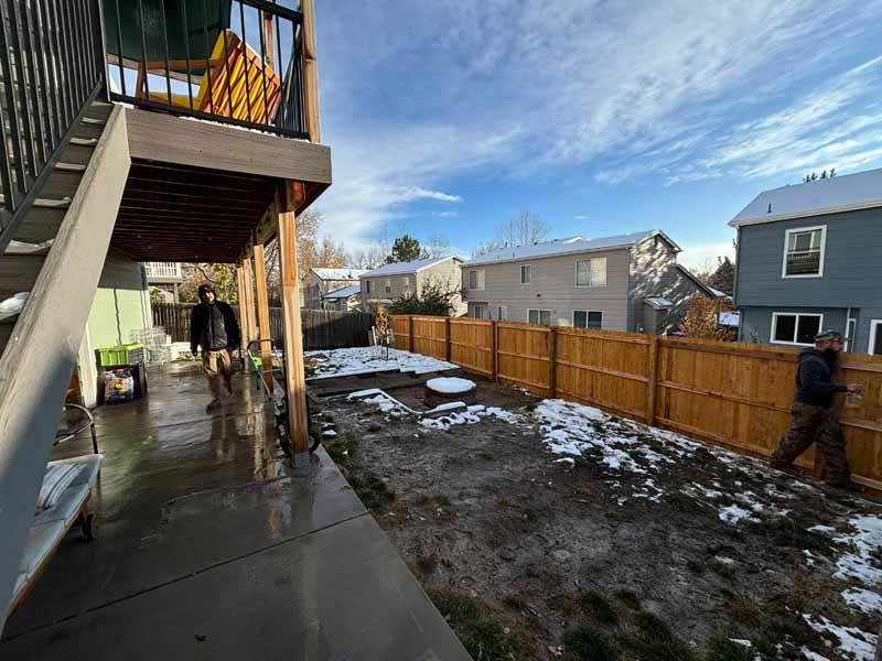 Two people work on a wooden fence in a partially snow-covered backyard beneath a deck, with houses in the background.