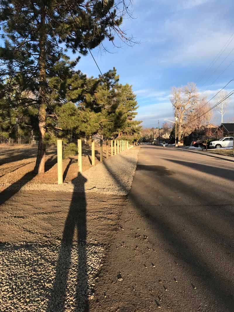 A long row of wooden posts along a dirt path next to a paved road on a sunny day, with a long shadow cast in the foreground.