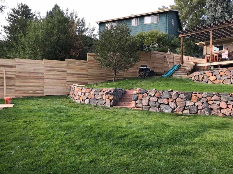 A grassy backyard with a terraced stone retaining wall, wooden stairs, and a tall horizontal-slat wooden privacy fence.