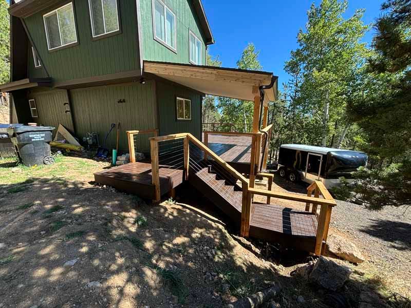 A green, two-story house features a wooden deck and staircase leading down to a dirt yard with a trailer in the background.