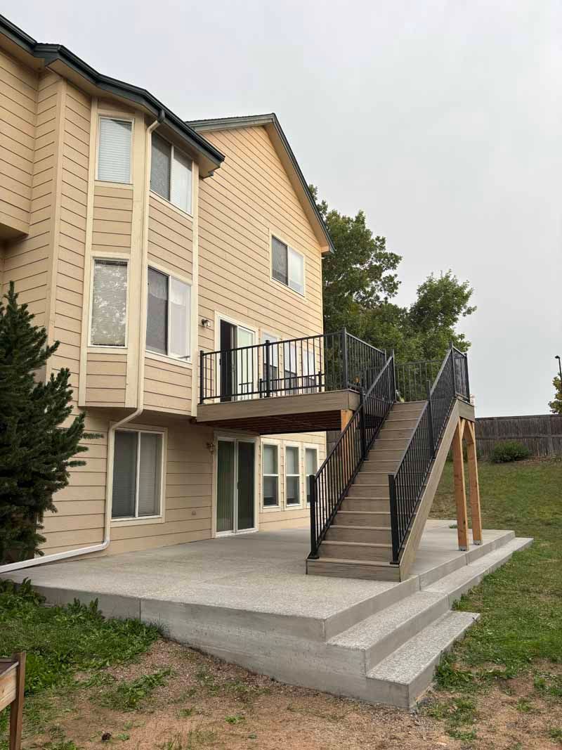 A beige multi-story house with a rear wooden deck and stairs leading down to a tiered concrete patio in a grassy yard.