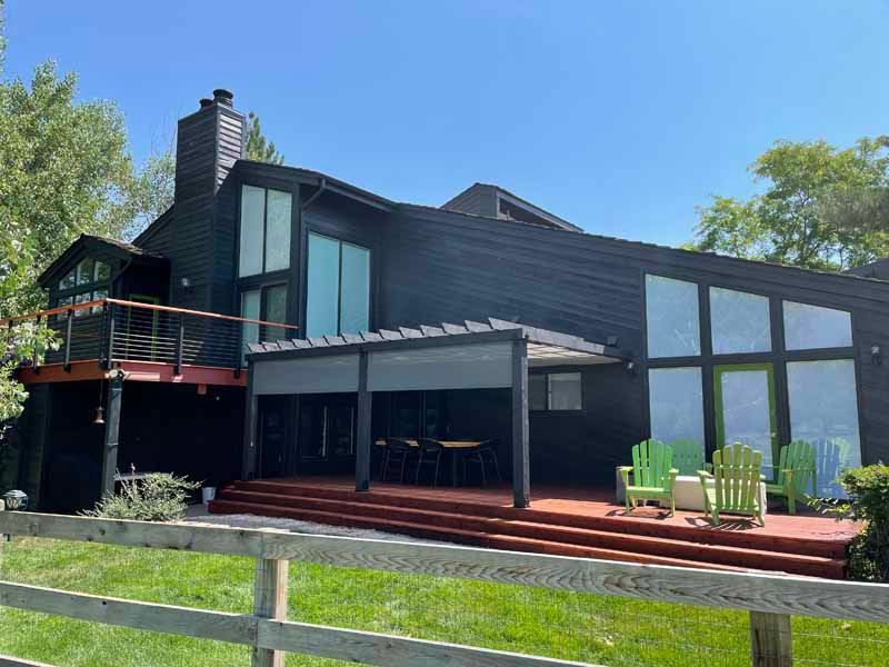 A modern black house with a red wood deck, pergola, and patio chairs, set behind a wooden fence on a sunny day.