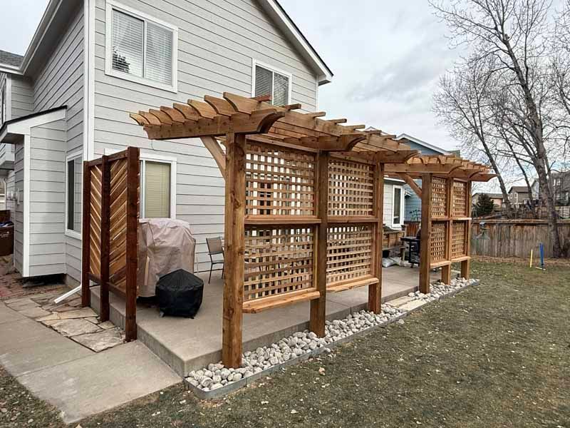 A multi-section wooden pergola with lattice privacy screens on a backyard concrete patio next to a gray house.