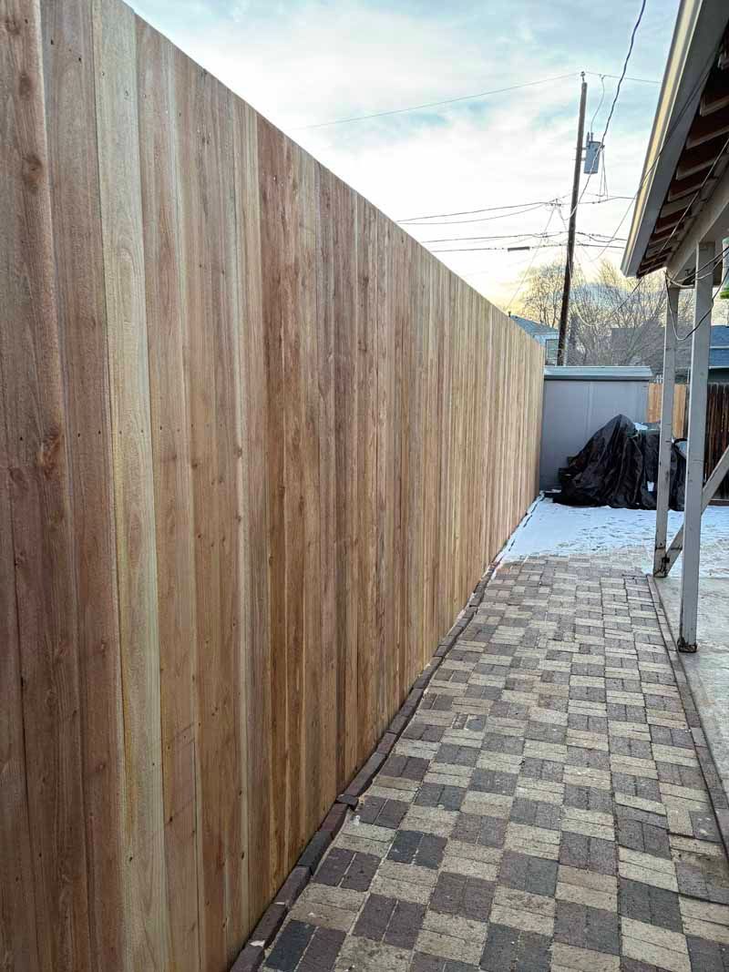 A long, light-brown wood privacy fence running along a checkerboard-patterned brick walkway outdoors in winter.