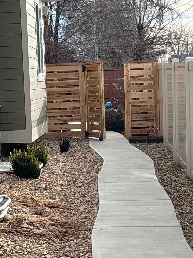 A concrete pathway leads to a wooden gate between two fences alongside the side of a house on a gravel-covered yard.
