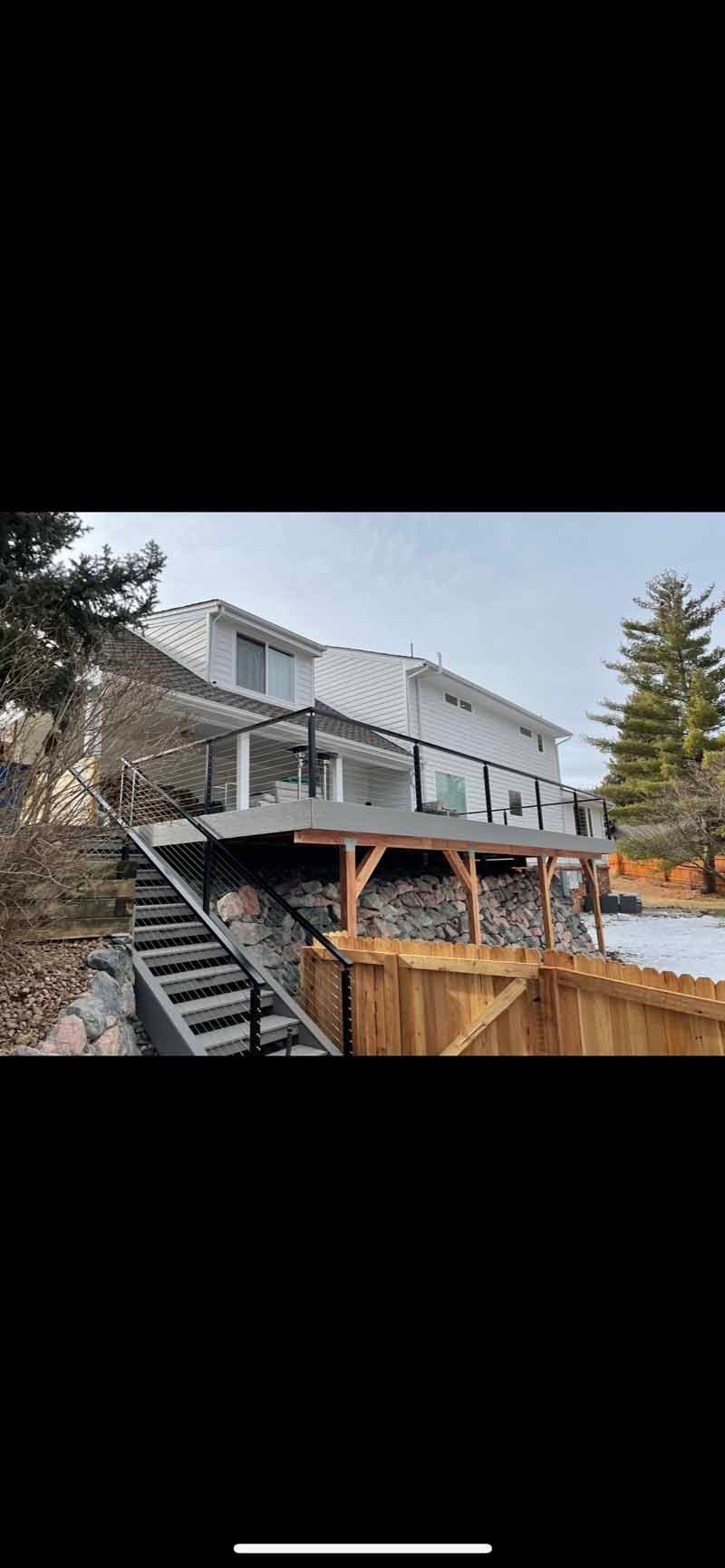 A white two-story house with a raised deck, black metal railings, and a wooden staircase leading down to a fenced yard.