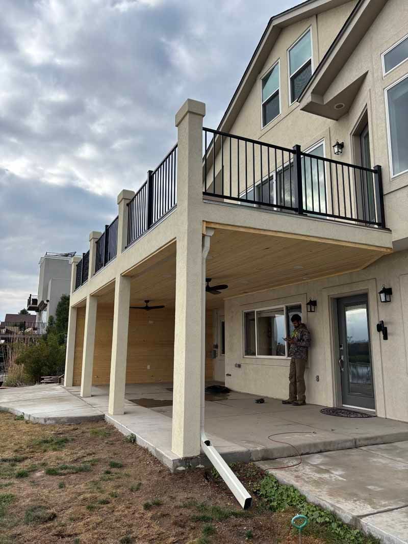 A two-story tan stucco house with a large upper-level deck supported by four columns above a lower concrete patio.