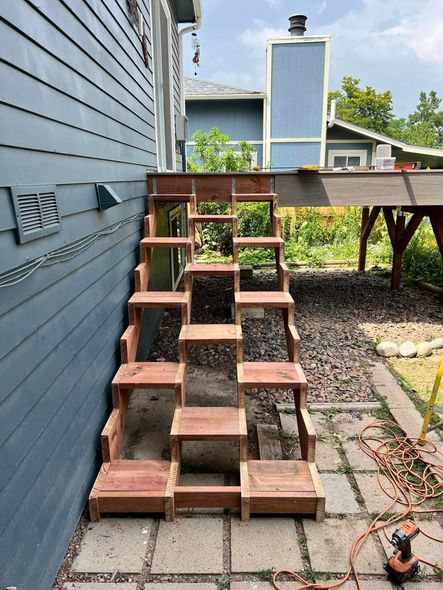 A wooden, split-level staircase built alongside a blue house leading up to a wooden deck outdoors.
