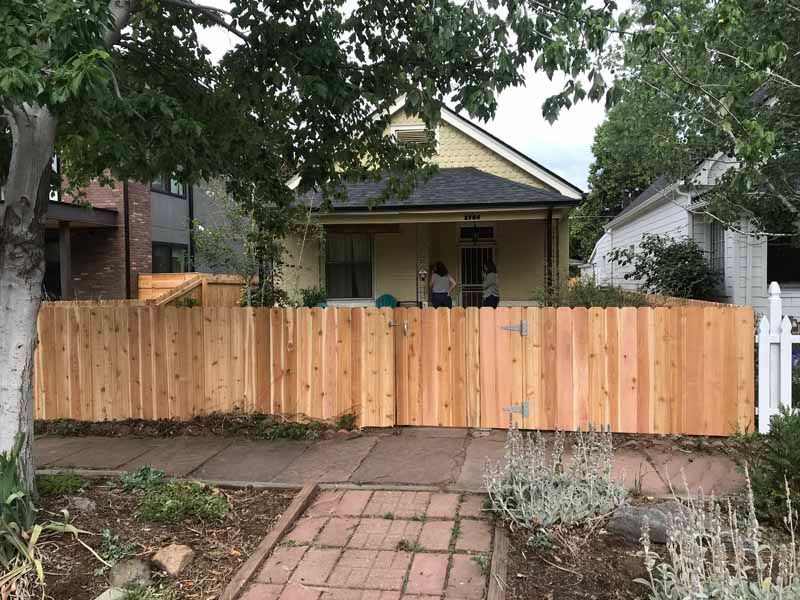 A new light-wood fence stands in front of a yellow house, with a brick path leading to the gate.
