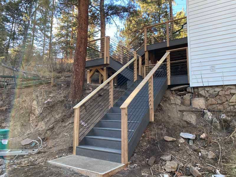 Gray outdoor stairs with wooden railings lead up a hillside to a deck attached to the side of a house.