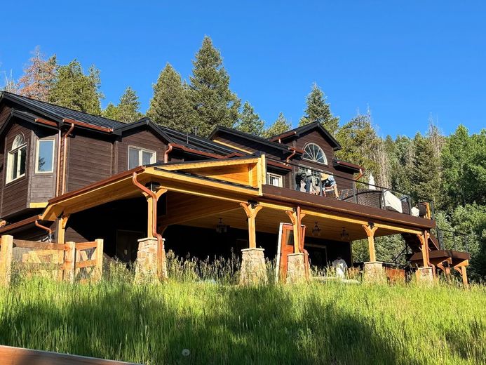 A large, dark-wood cabin with a spacious covered deck featuring stone pillar supports, set against a forest and blue sky.