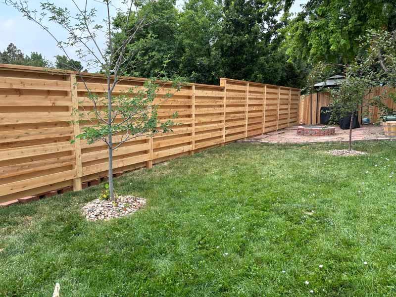 A wooden horizontal-slat fence runs along a green lawn in a backyard, with trees and a fire pit area in the background.