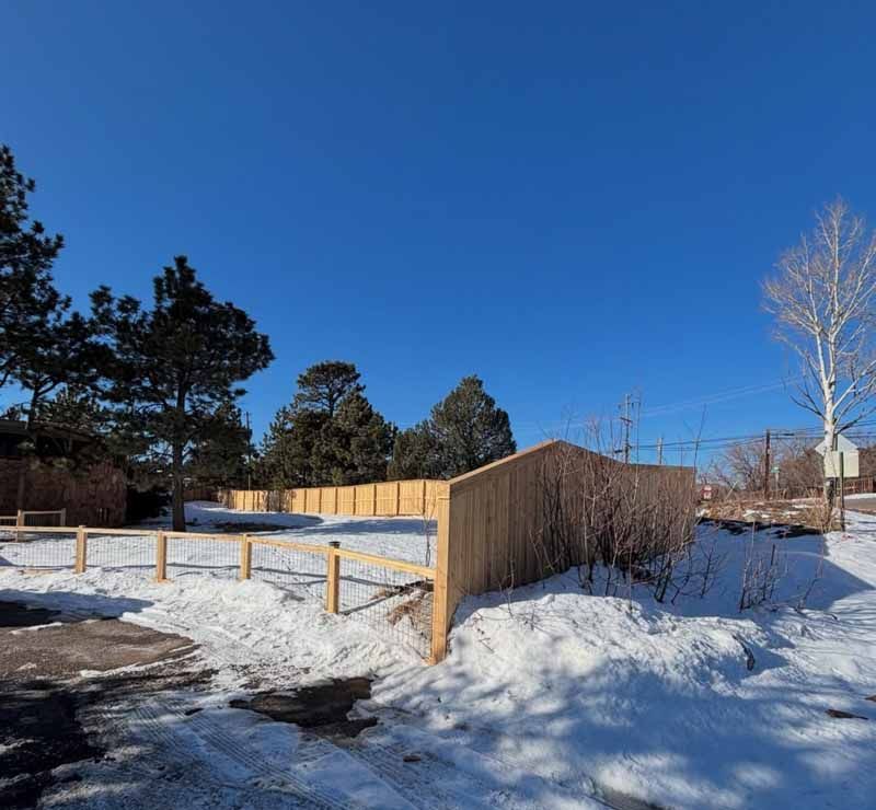 A tall, wooden privacy fence stands in a snowy, sunlit outdoor area near trees under a clear blue sky.