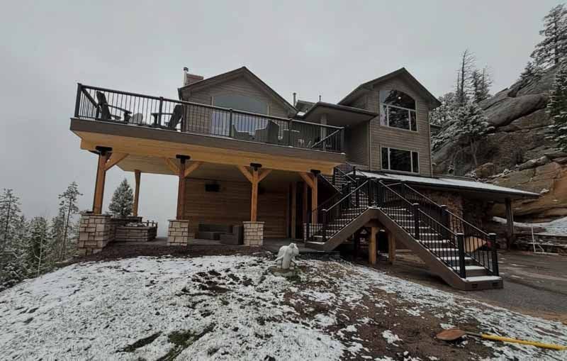A multi-story mountain home with a large wooden deck and outdoor staircase, set against a rocky, snow-covered slope.