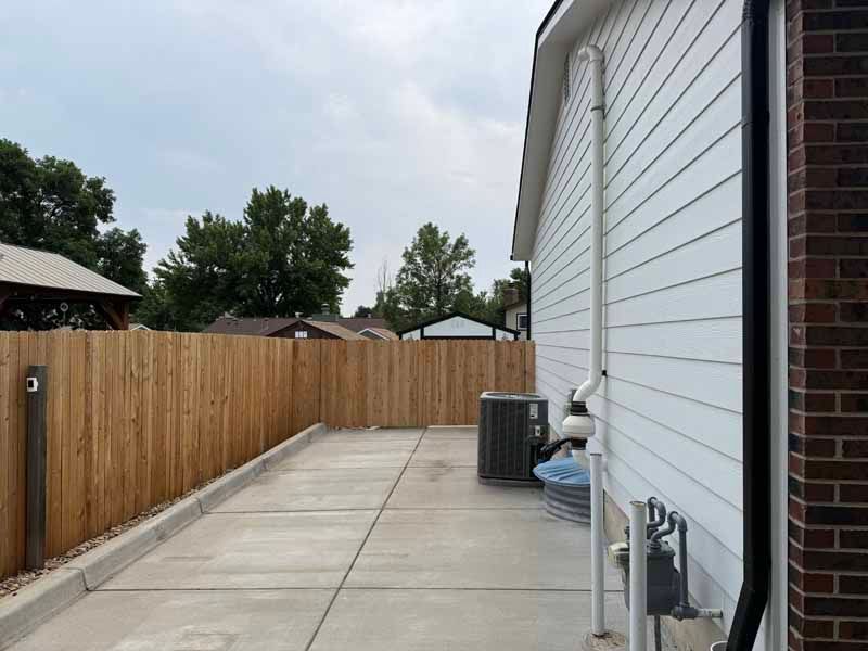 A concrete side yard with white siding, a metal fence, and mechanical equipment alongside a brick corner of a house.