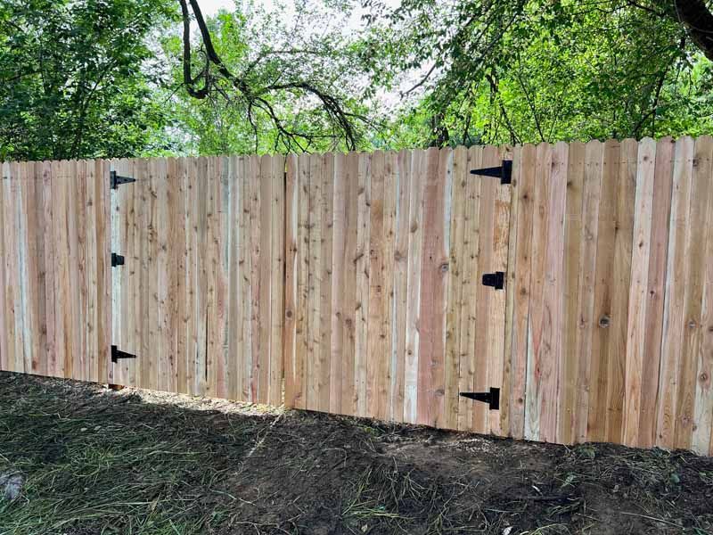 A wooden fence with two integrated gates featuring black T-hinges, set against a background of green trees.