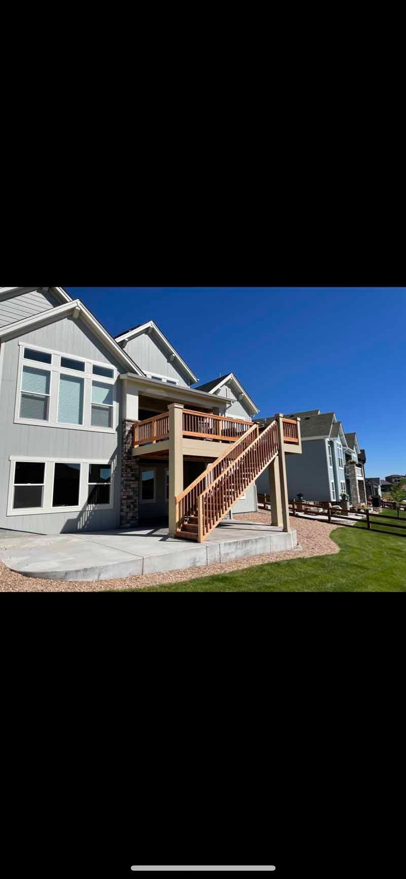 A two-story light gray house with a wooden deck, a staircase leading to a concrete patio, and a clear blue sky.