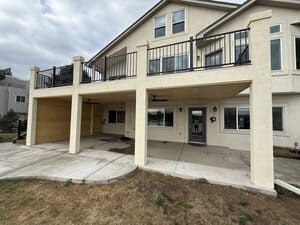 A beige two-story house with a large upper deck and a covered patio area with a concrete floor.