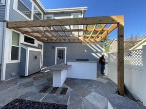 A person stands in a gray-toned patio with a wooden pergola over a white outdoor bar and stone tile flooring.