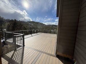 A spacious, light-colored wooden deck with black railings overlooking a forest on a sunny day with partly cloudy skies.