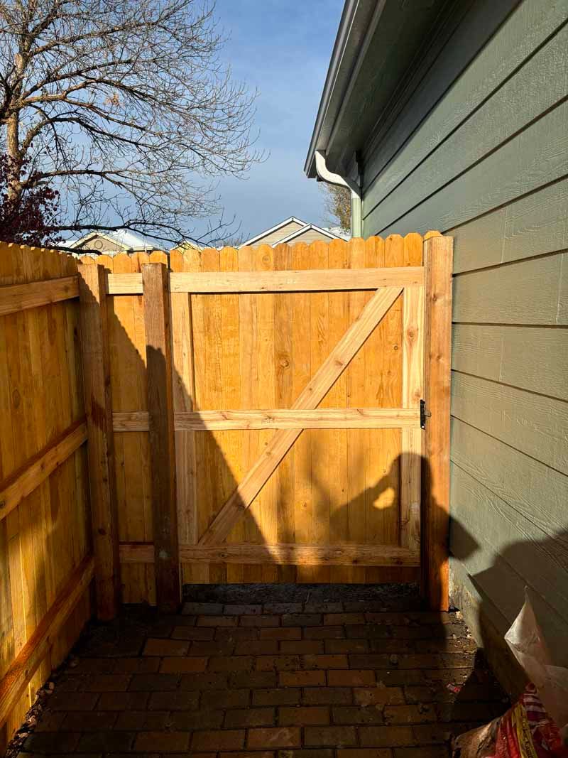 A new wooden gate installed between a brick patio and the siding of a house.