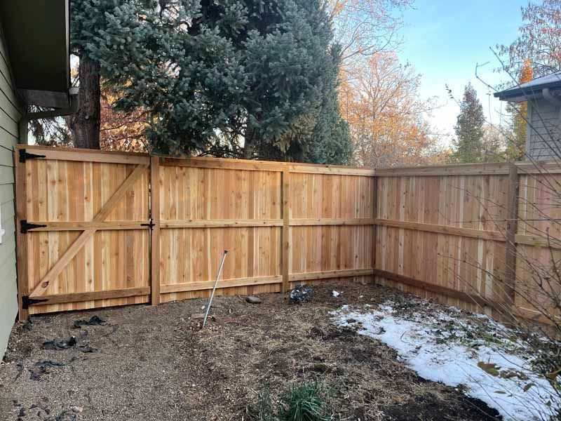 A new cedar wood privacy fence with a gate, located in a yard with sparse grass and patches of snow.