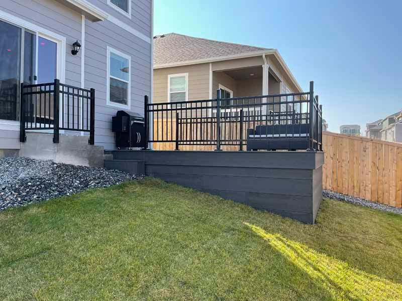 A backyard view showing a gray deck with black metal railings, a stone area, and a grassy slope on a sunny day.
