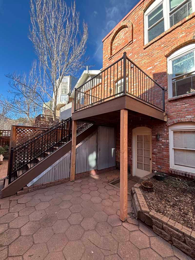A brick building with an elevated wooden deck and staircase leading to a patio area paved with hexagonal stones.