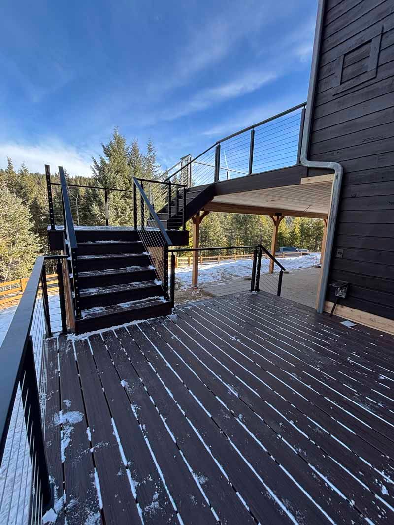 A dark wooden deck with modern railing leads to a staircase descending to a snowy, tree-lined yard under a blue sky.