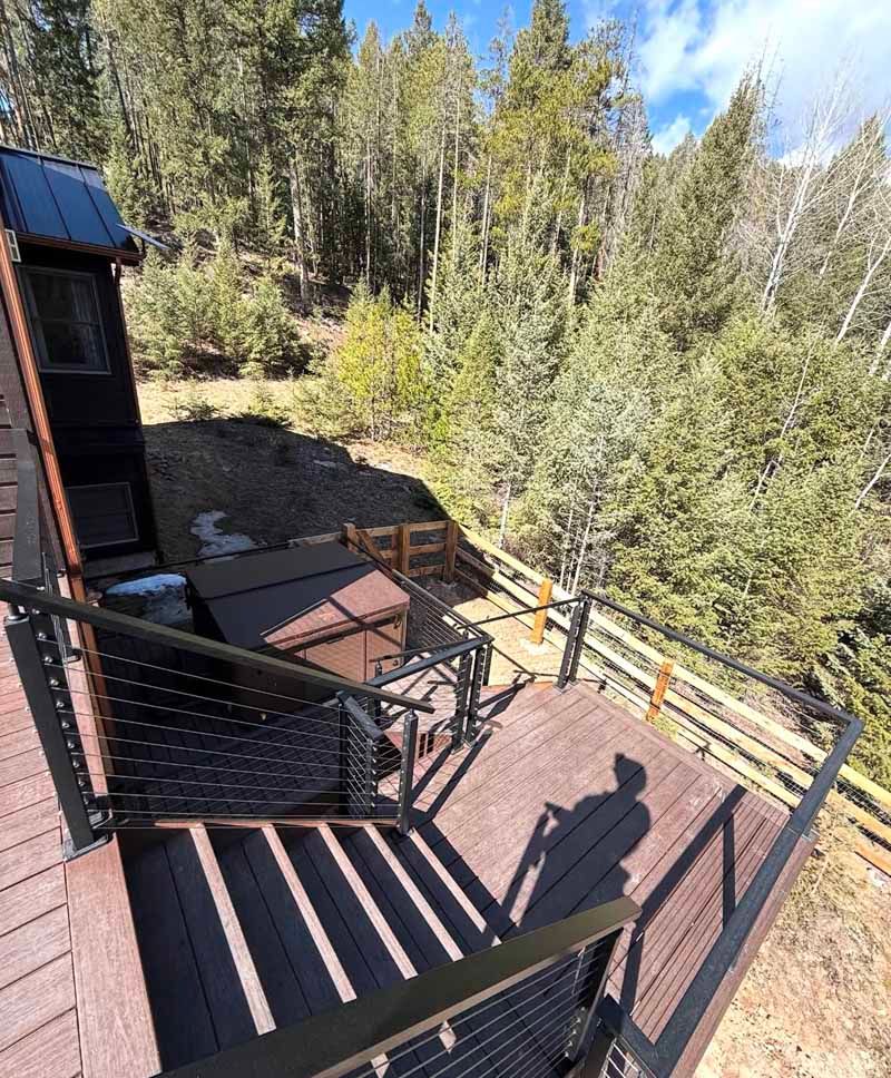 An elevated wooden deck with cable railings overlooks a forest, featuring a square hot tub and a house exterior nearby.