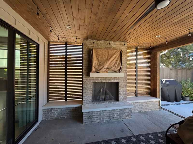 A covered outdoor patio featuring a central stone fireplace, horizontal wooden slats, and string lights on a wood ceiling.