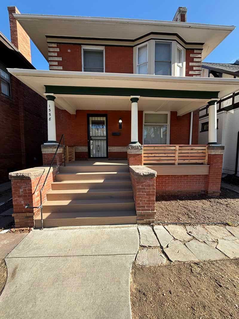 A two-story red brick house with a wide front porch, concrete steps, white columns, and a wooden porch railing.