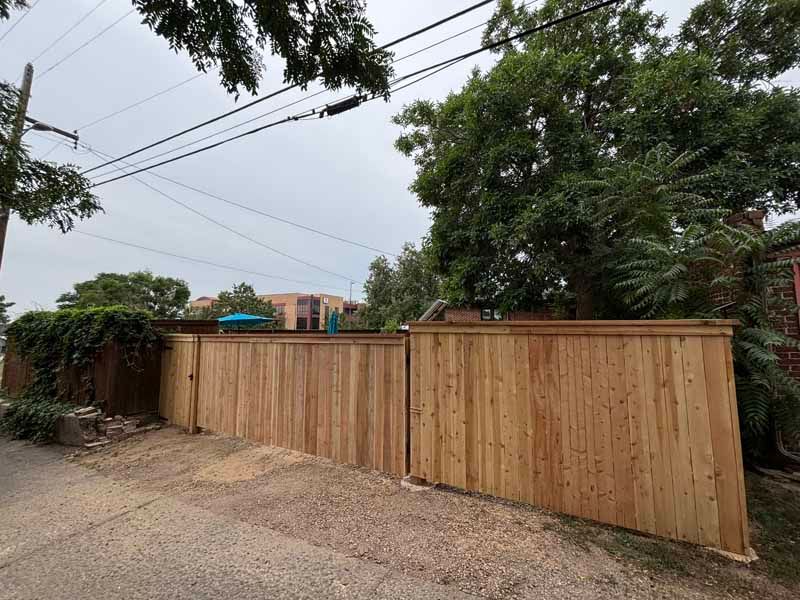 A newly installed natural wood fence lines a gravel path, with a large tree and building visible in the background.