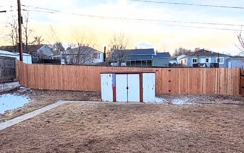 A wooden privacy fence crosses a backyard, with a small, white, flat-roofed storage shed centered against it.