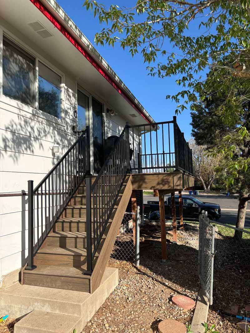A wooden deck with black railings and stairs leading to an elevated door on a white house exterior under a clear blue sky.