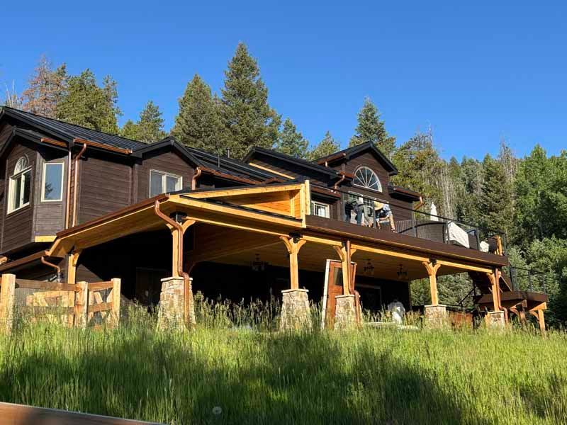 A dark brown multi-story wooden house with a spacious covered porch on stone pillars, set against a forest and blue sky.